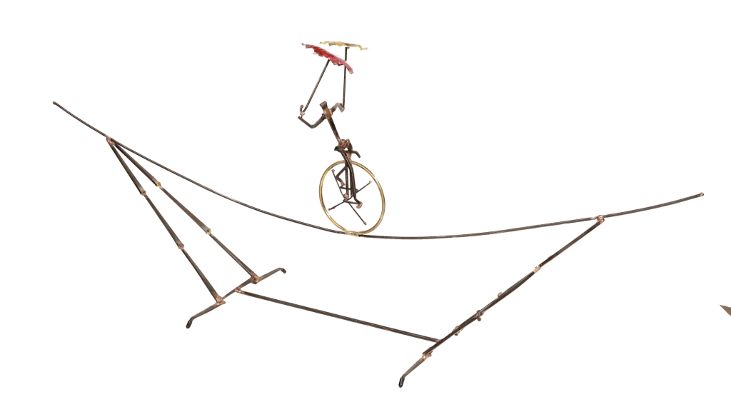 Freestanding metal sculpture depicting an acrobatic unicyclist, balancing with two umbrellas, one brass and one copper, with the unicycle supported on an intricate metal tightrope structure.