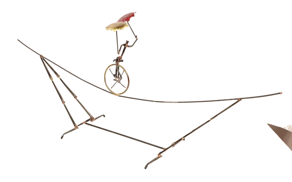 Freestanding metal sculpture depicting an acrobatic unicyclist, balancing with two umbrellas, one brass and one copper, with the unicycle supported on an intricate metal tightrope structure.