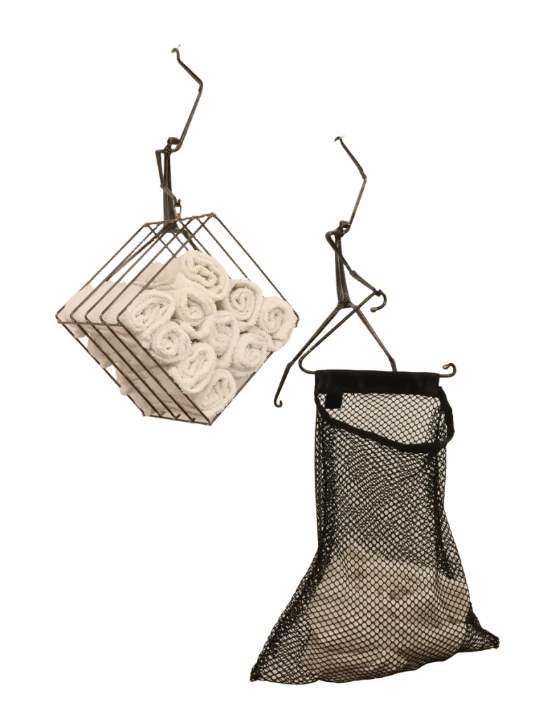 Wall-mounted bathroom storage unit metal sculpture with rolled white towels in a steel wire basket, a black polyester mesh removable laundry bag for soiled hand towels hanging below from a steel hanger and incorporating a dock side theme with a decorative steel dragonfly, copper and brass reed accents and steel birds in flight from a brass dock pier.