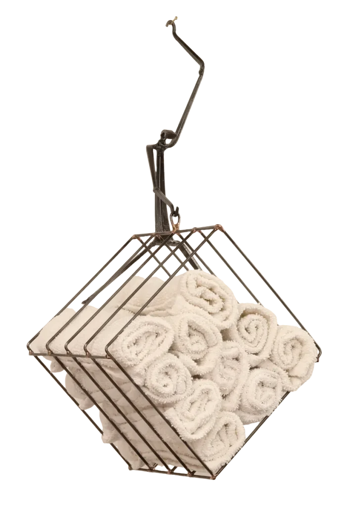 Wall-mounted bathroom storage unit metal sculpture with rolled white towels in a steel wire basket, a black polyester mesh removable laundry bag for soiled hand towels hanging below from a steel hanger and incorporating a dock side theme with a decorative steel dragonfly, copper and brass reed accents and steel birds in flight from a brass dock pier.