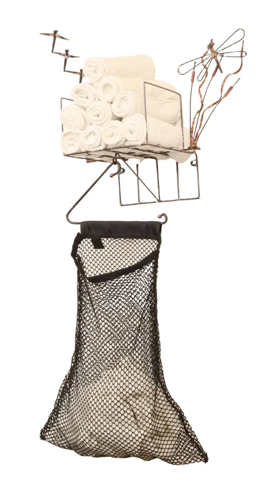 Wall-mounted bathroom storage unit metal sculpture with rolled white towels in a steel wire basket, a black polyester mesh removable laundry bag for soiled hand towels hanging below from a steel hanger and incorporating a dock side theme with a decorative steel dragonfly, copper and brass reed accents and steel birds in flight from a brass dock pier.
