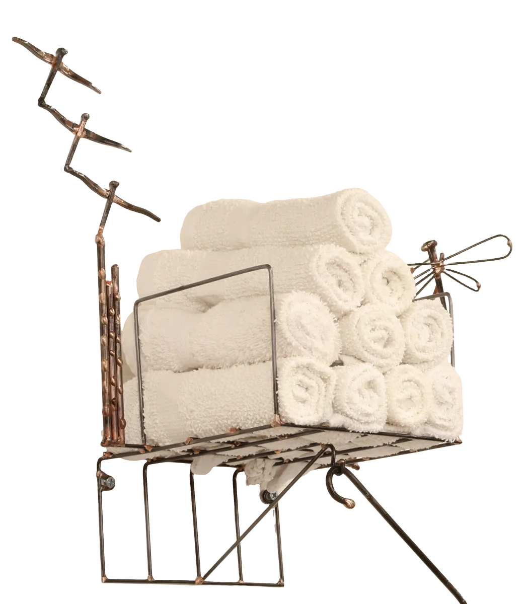 Wall-mounted bathroom storage unit metal sculpture with rolled white towels in a steel wire basket, a black polyester mesh removable laundry bag for soiled hand towels hanging below from a steel hanger and incorporating a dock side theme with a decorative steel dragonfly, copper and brass reed accents and steel birds in flight from a brass dock pier.
