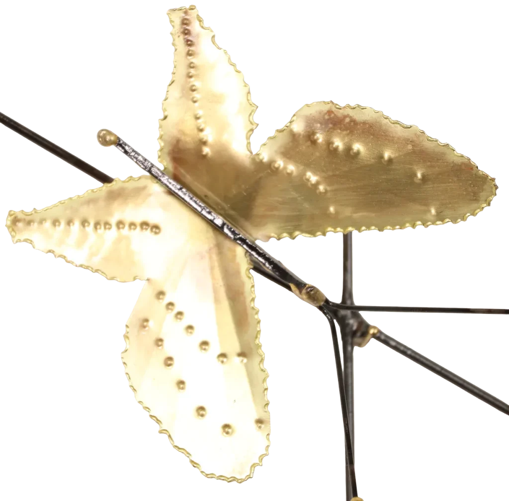 Decorative metal wall art depicting a trellis-like structure adorned with brass leaves, deep red copper roses of three different sizes, a brass butterfly and steel wire hummingbird feeding from one of the copper roses. A small spider-like figure is visible near the upper left.