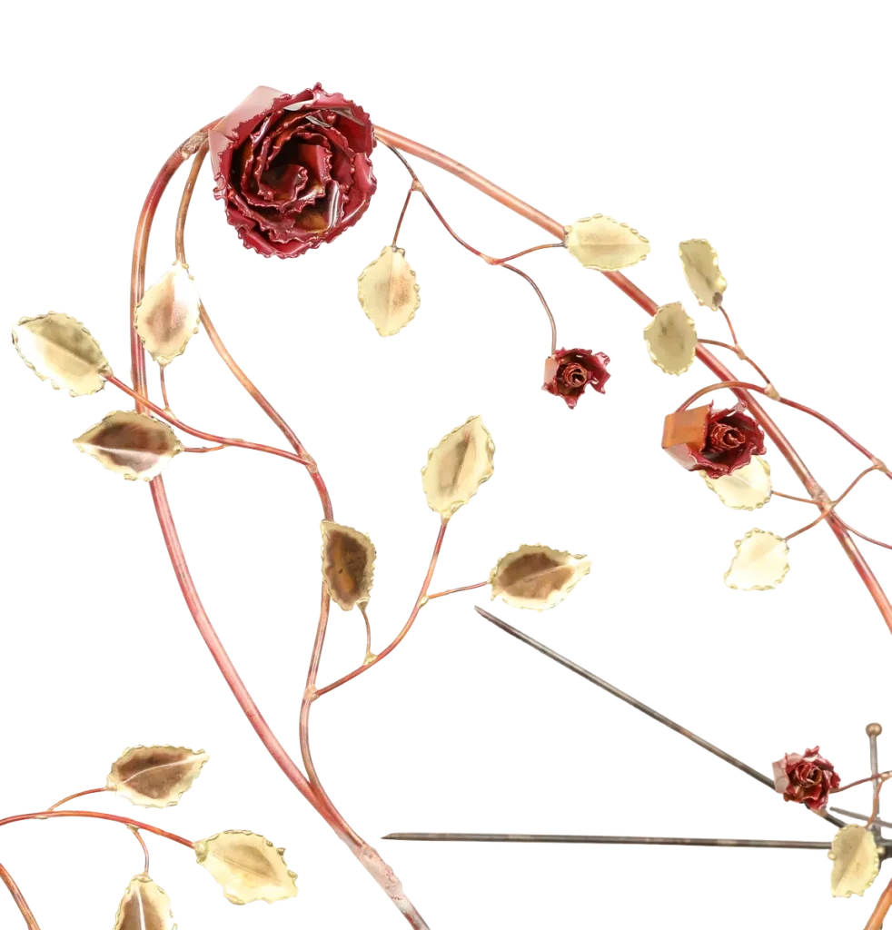 Decorative metal wall art depicting a trellis-like structure adorned with brass leaves, deep red copper roses of three different sizes, a brass butterfly and steel wire hummingbird feeding from one of the copper roses. A small spider-like figure is visible near the upper left.