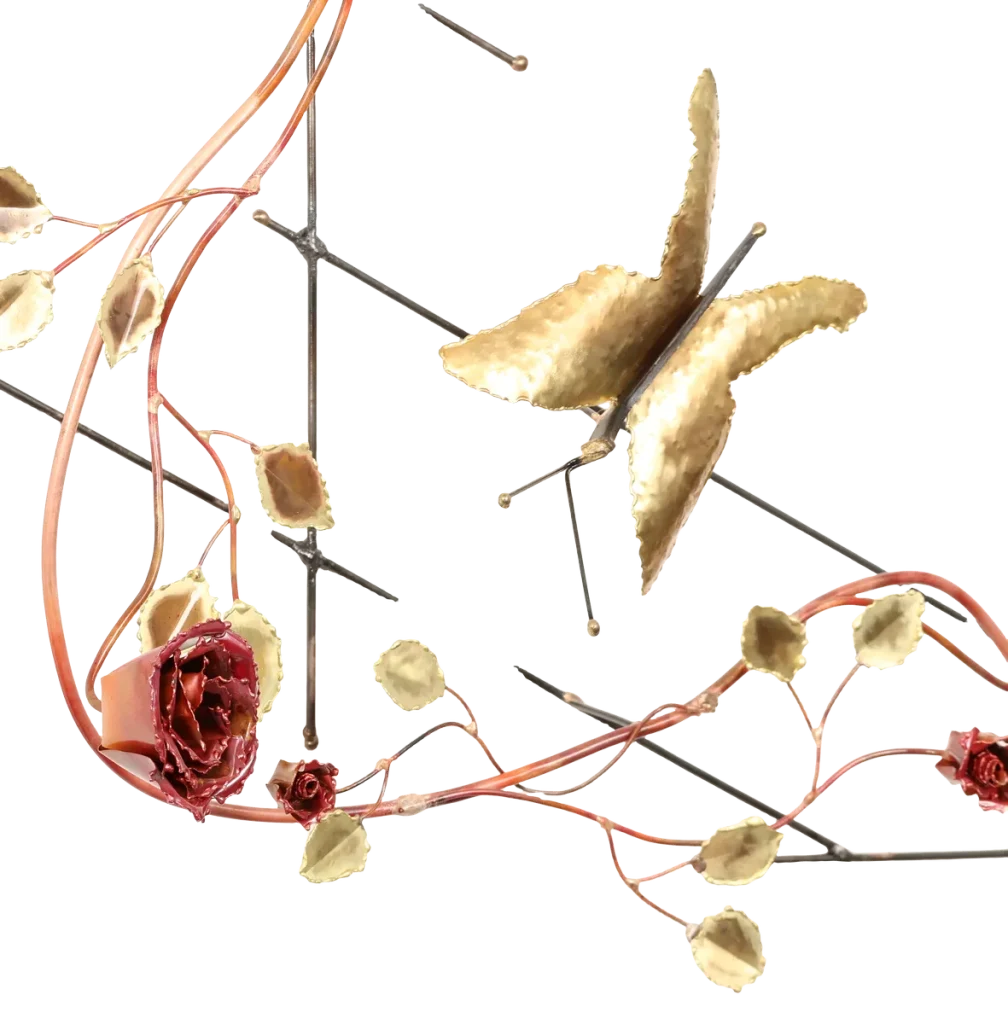 Decorative metal wall art depicting a trellis-like structure adorned with brass leaves, deep red copper roses of three different sizes, a brass butterfly and steel wire hummingbird feeding from one of the copper roses. A small spider-like figure is visible near the upper left.