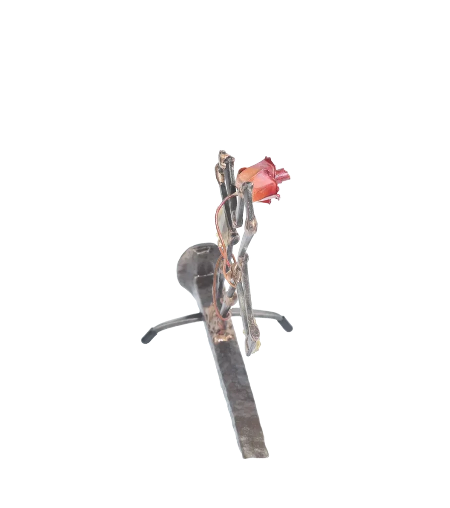 Freestanding metal sculpture depicting a broken cross made of cut steel nails and mounted on a railroad spike with a copper rose and vine with brass leaves, growing out of the break in the cross. The broken cross and rose is symbolic of the resurrection of Jesus after crucifixion.