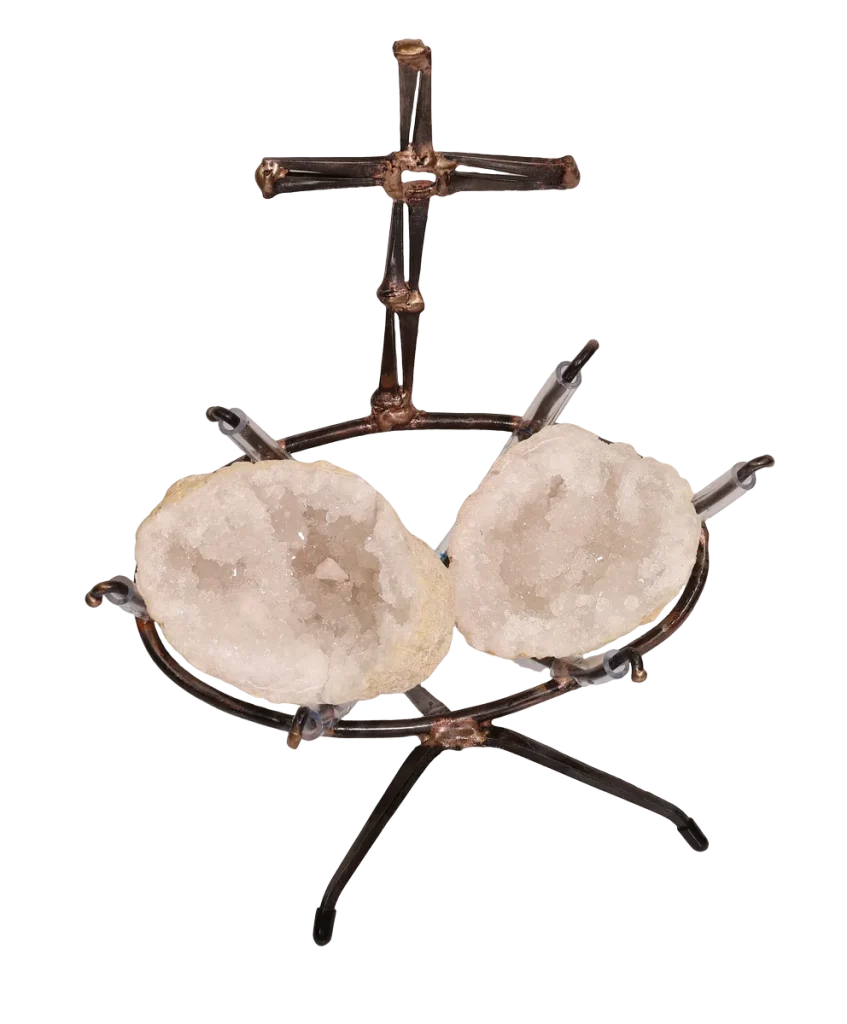 A handmade metal sculpture featuring a cross made of steel cut nails and rising above two white geode halves held within a steel clear vinyl padded frame resting on a tripod base.