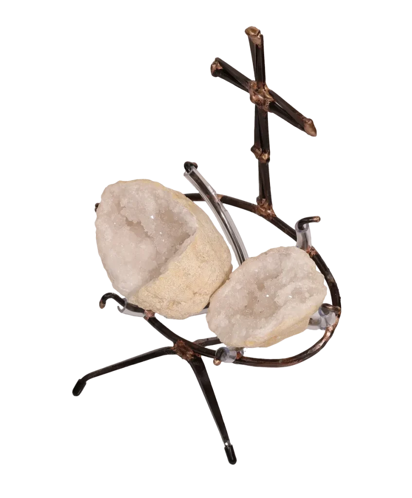A handmade metal sculpture featuring a cross made of steel cut nails and rising above two white geode halves held within a steel clear vinyl padded frame resting on a tripod base.