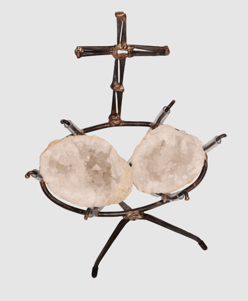 A handmade metal sculpture featuring a cross made of steel cut nails and rising above two white geode halves held within a steel clear vinyl padded frame resting on a tripod base.